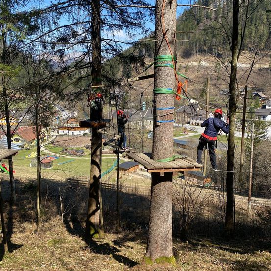 Drei Personen navigieren einen Baumkletterkurs mit Sicherheitsseilen und Gurten, in einem üppig grünen Wald mit Häusern und Bergen im Hintergrund.