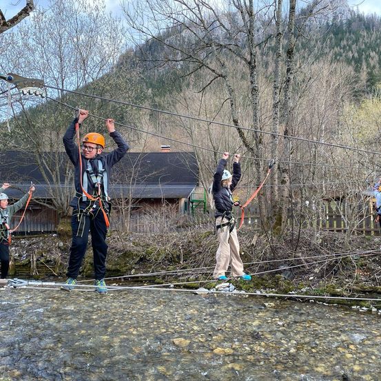 Drei Personen gehen auf einem Drahtseil über einem Bach, mit Bergen im Hintergrund.