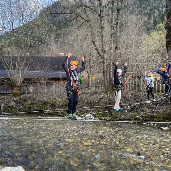 Fünf Personen überqueren eine Hängebrücke über einem Fluss, tragen Sicherheitsausrüstung und Helme. Die Brücke wird von Seilen getragen und befindet sich in einem Gebirgsgebiet mit einem Holzgebäude im Hintergrund.