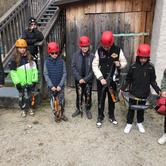 Fünf Kinder mit Helmen und Gurten stehen vor einer Holztür. Dahinter steht eine Frau auf der Treppe und ein Mann sitzt auf einer Bank.