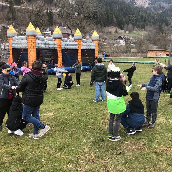 Eine Gruppe von Kindern und Erwachsenen versammelt sich auf einem Rasenfeld. Einige springen und spielen in einer aufblasbaren Burg, während andere zuschauen oder herumstehen. Im Hintergrund befinden sich Häuser und Bäume.