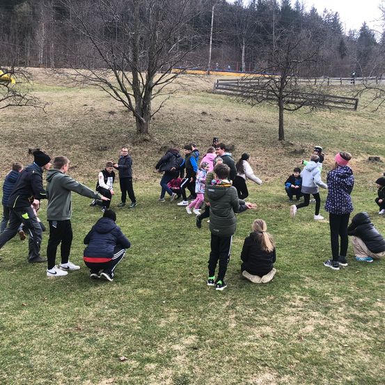 Eine Gruppe von Menschen in Winterkleidung versammelt sich auf einem Grasfeld, einige stehen, einige sitzen und einige sind in Bewegung, mit einem Waldhintergrund und einem Holzzaun in der Ferne.