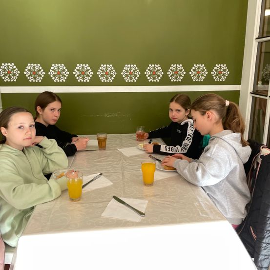 Fünf Mädchen sitzen an einem Tisch mit weißer Tischdecke und trinken Orangensaft. Sie sitzen in einem Raum mit grünen Wänden und dekorativen Mustern.