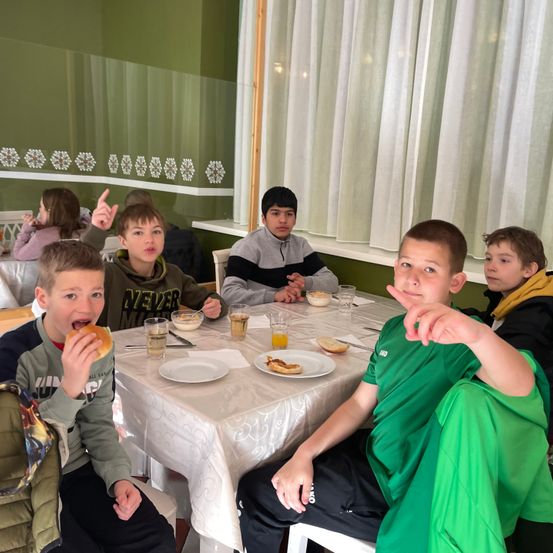 Eine Gruppe von Jungen sitzt an einem Tisch in einem Raum, einige essen und andere posieren für ein Foto. Eines Mädchen im Hintergrund isst ebenfalls.