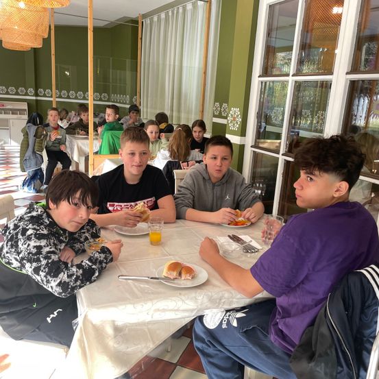 Vier Jungen sitzen an einem Tisch in einem Restaurant und essen. Dahinter sitzen weitere Kinder, die ebenfalls essen.
