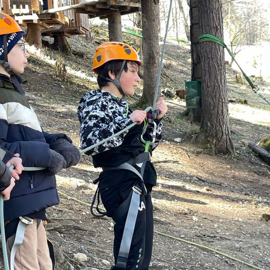 Zwei Kinder klettern im Freien und tragen orangefarbene Helme und Gurte. Ein Kind hält das Seil fest umklammert. Im Hintergrund befindet sich eine Holzplattform und Bäume. Der Boden ist mit Erde und kleinen Steinen bedeckt.