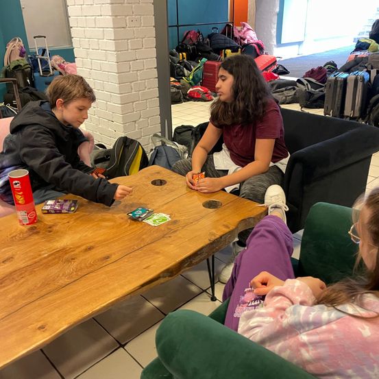 Zwei junge Leute spielen ein Kartenspiel an einem Holztisch. Ein Mädchen mit einer roten Bluse und Jeans sitzt auf einem Stuhl mit überkreuzten Beinen. Ein Junge in einer schwarzen Jacke sitzt auf einem rosa Stuhl.