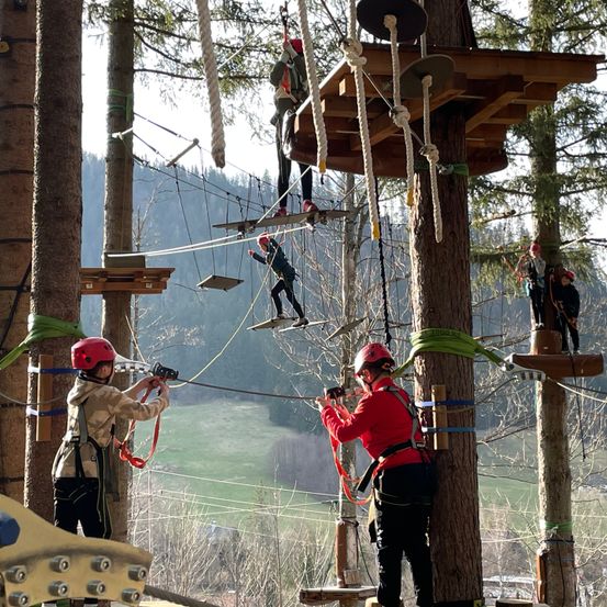 Mehrere Personen mit Helmen klettern auf und ab der Seilbrücke. Sie halten sich an den Seilen fest und tragen Gurte.