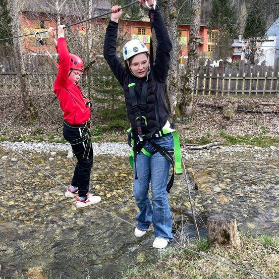 Zwei junge Frauen gehen auf einem Seil über einen Fluss, mit Helmen und Gurten gesichert.