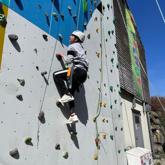 Ein Junge mit weißem Helm und Turnschuhen klettert an einer Kletterwand mit den Händen an den Griffen.