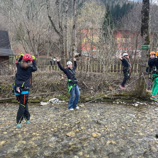 Vier Personen in Helmen und Gurten gehen auf einem Seil, das über einen Fluss gespannt ist.