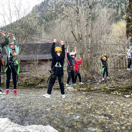 Eine Gruppe von Menschen in Helmen überquert eine Hängebrücke über einen Fluss mit Bäumen auf beiden Seiten.
