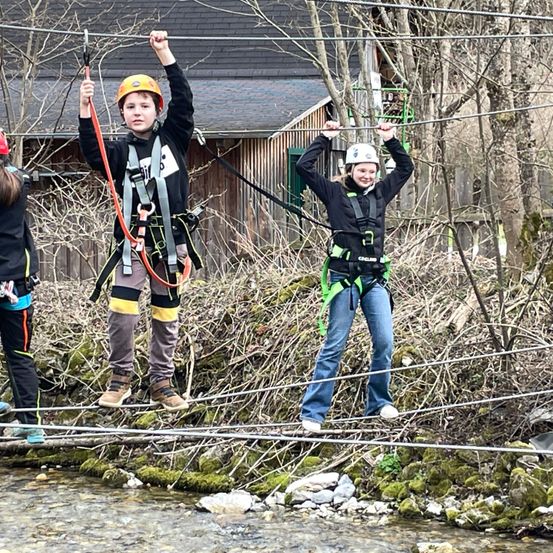 Drei Kinder tragen Helme und Gurte und gehen auf einem Seil, das über einem Fluss gespannt ist. Dahinter befindet sich ein Holzgebäude mit einem schwarzen Dach.
