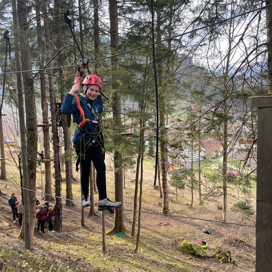 Ein Mädchen in einer blauen Jacke, schwarzen Hosen und einem roten Helm fährt eine Seilbahn über dem Boden in einem Wald.