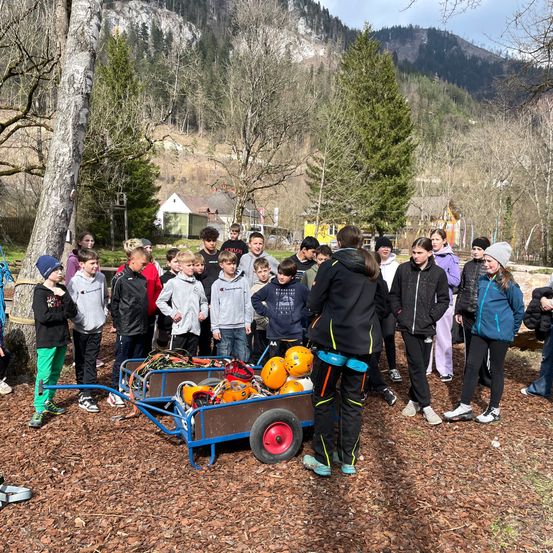 Eine Gruppe junger Menschen steht um einen blauen Schubkarren voller orangefarbener Sicherheitshelme herum. Sie tragen Jacken und Turnschuhe, und einige von ihnen tragen Mützen. Sie stehen in einem Außenbereich mit Bäumen und Bergen in der Ferne.