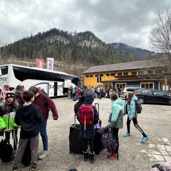 Eine Gruppe von Kindern und Erwachsenen versammelt sich um einen Bus und ein Gebäude. Einige gehen mit ihrem Gepäck. Ein Berg und Bäume sind im Hintergrund.
