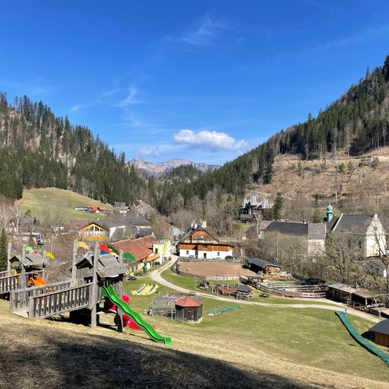 Ein Blick von oben auf ein kleines Dorf mit vielen Häusern und einem Spielplatz im Vordergrund. Das Dorf ist von Bäumen und Bergen umgeben.