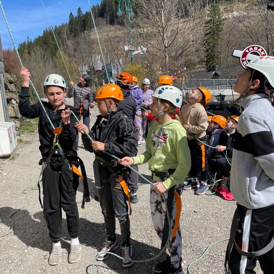 Gruppe von Kindern in Helmen klettert an einem Seil mit angehängten Seilen, die auf einem Schotterweg stehen, mit Bäumen und Bergen im Hintergrund.