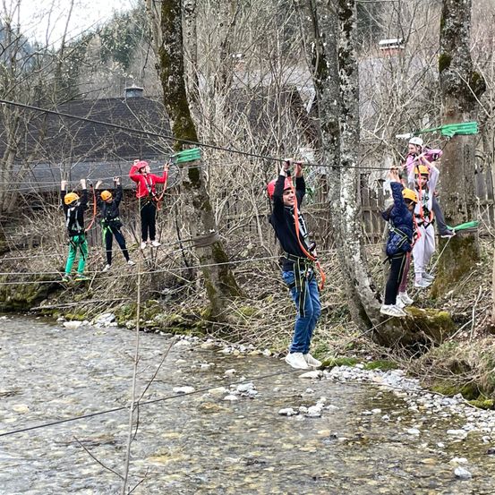 Eine Gruppe von Menschen zippt auf einem Seil über einen Fluss, mit Helmen und Gurten. Die Seilbahn ist zwischen Bäumen auf beiden Seiten des Flusses aufgehängt.