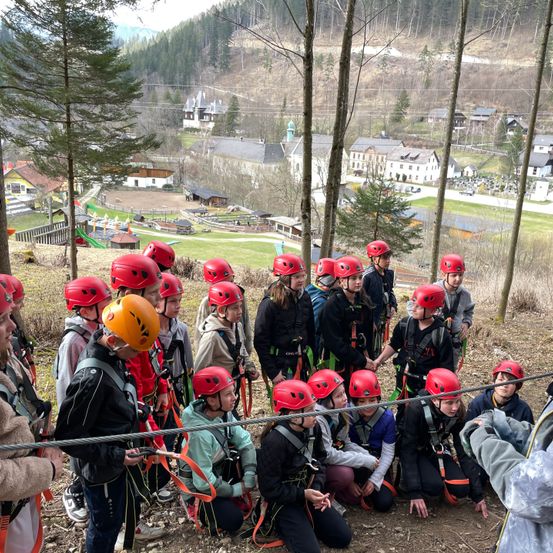 Eine Gruppe von Menschen steht auf einem Erdfeld und trägt rote Helme und Gurte. Sie scheinen sich auf ein Abenteuer vorzubereiten. Das Gebiet ist von Bäumen und Gras umgeben. In der Ferne befindet sich ein Gebäude mit einem kuppelförmigen Dach.