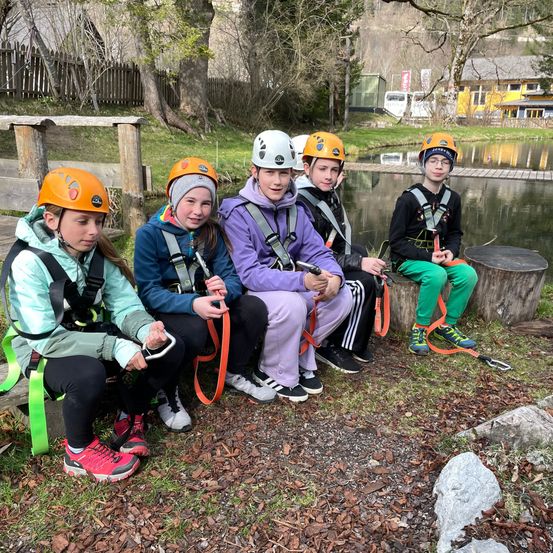 Eine Gruppe von Kindern mit Helmen sitzt auf einem Baumstumpf in der Nähe eines Sees.