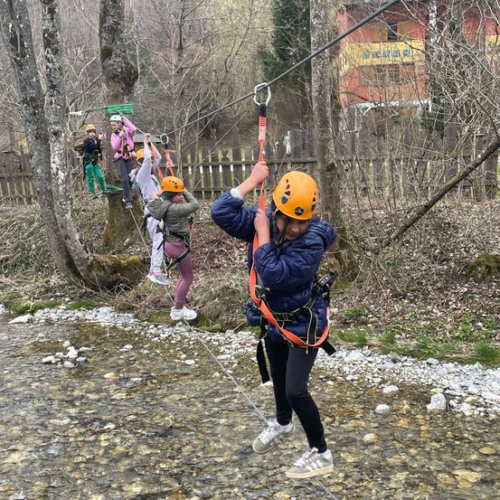 Eine Gruppe von Menschen in Helmen und Sicherheitsgurten fährt mit einer Seilbahn über einen Bach. Die Seilbahn ist an einem Baum auf der anderen Seite befestigt.