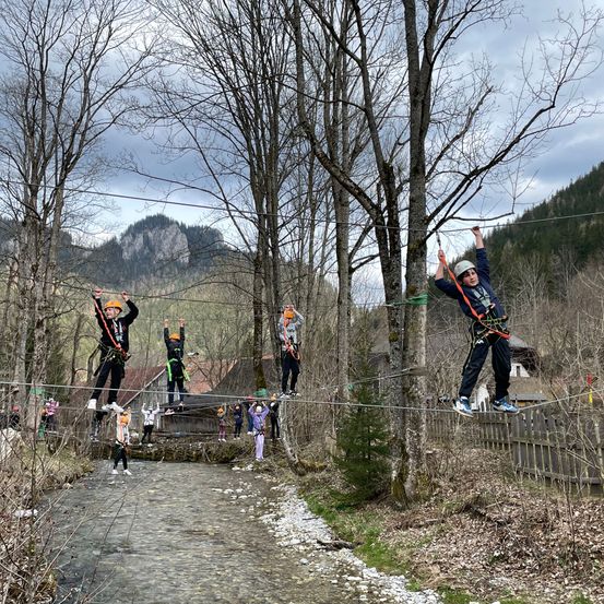 Eine Gruppe von Menschen überquert ein Seil, das über einem Fluss gespannt ist, mit Helmen und Gurten. Sie stehen auf den Seilen, einige halten sich an den Seilen fest.