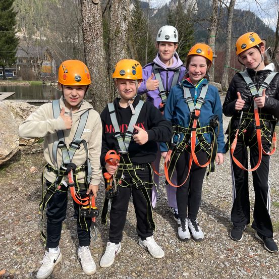 Fünf junge Leute stehen nebeneinander und tragen orangefarbene Helme und Gurte. Sie lächeln und geben ein Daumen hoch. Dahinter sind Bäume und ein Gewässer.