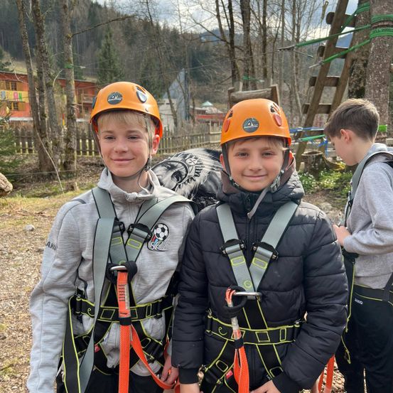 Zwei Jungen mit orangefarbenen Helmen und Gurten lächeln in die Kamera, einer in einem grauen Sweatshirt und der andere in einer schwarzen Jacke, mit einem Jungen, der rechts neben ihnen steht, und einer Holzleiter dahinter.