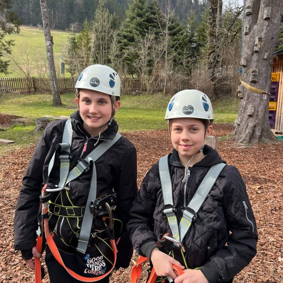 Zwei Kinder in Sicherheitsgurten stehen nebeneinander und lächeln. Sie tragen weiße Helme.