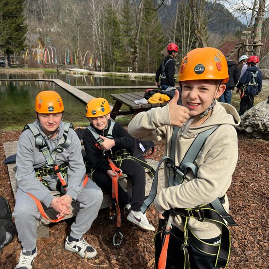 Drei junge Jungen in orangefarbenen Helmen und Gurten sitzen auf einer Holzbank, einer von ihnen gibt einen Daumen hoch. Dahinter steht ein Holztisch und eine Person, während in der Ferne ein See mit einem Holzsteg und Bäumen zu sehen ist.