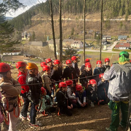 Eine Gruppe junger Menschen mit roten Helmen versammelt sich im Wald, mit Bäumen und Bergen im Hintergrund.