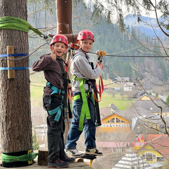 Zwei junge Jungen in Helmen und Gurten klettern einen Baum hinauf. Sie stehen auf einer Plattform, die mit Seilen und Drähten am Baum befestigt ist. Sie lächeln und posieren für ein Foto. Dahinter ist ein Blick auf ein Dorf mit Häusern und Bergen.
