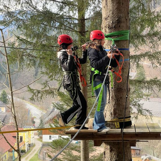 Zwei Frauen bereiten sich auf eine Klettertour auf einem an einem Baum befestigten Plattform vor, tragen rote Helme und Gurte.