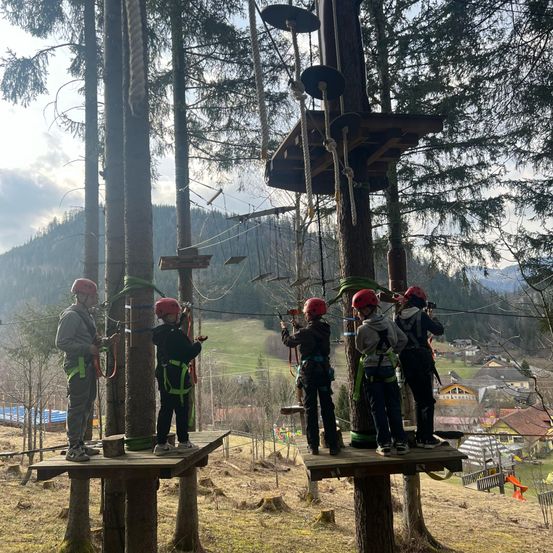 Fünf Kinder mit roten Helmen und Gurten stehen auf Plattformen in einem Wald und bereiten sich darauf vor, eine Seilbahn zu erklimmen. Im Hintergrund ist ein bergiges Gebiet mit Häusern zu sehen.