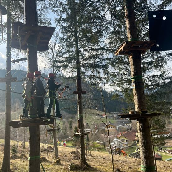 Eine Gruppe von Menschen in Helmen steht auf einer Plattform, hält sich an Seile, die mit Bäumen verbunden sind, in einem Außenbereich, wahrscheinlich in einem Wald, mit einem Dorf in der Ferne.