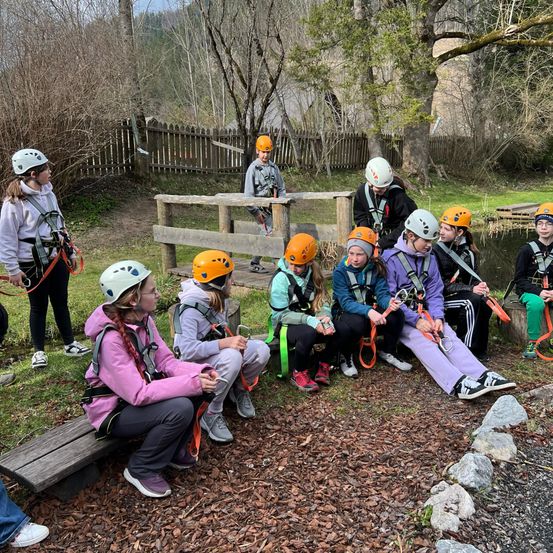 Eine Gruppe von Kindern mit Helmen sitzt auf einer Bank und einer steht bei einem Holzzaun.