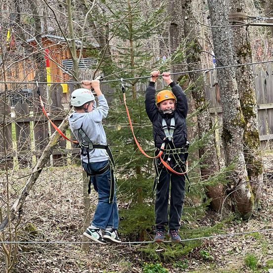 Zwei Kinder sind auf einer Zipline. Sie lächeln und halten sich an der Zipline fest.