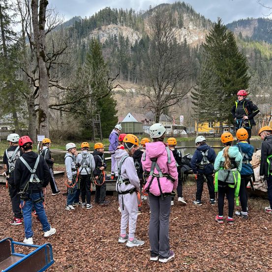 Eine Gruppe von Kindern steht in einem Kreis und trägt Sicherheitshelme und Gurte. Eine Person steht auf einem Felsen in der Mitte der Gruppe. Ein Berg ist im Hintergrund.