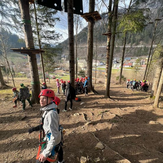 Eine Gruppe von Menschen geht in einem Wald mit Bäumen, Bergen und Häusern in der Ferne. Einige von ihnen tragen rote Helme und Gurte.