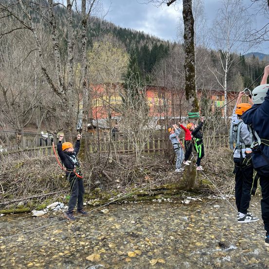 Eine Gruppe von Menschen macht Ziplining über einen Bach. Sie tragen alle Sicherheitsausrüstung, darunter Helme und Gurte.