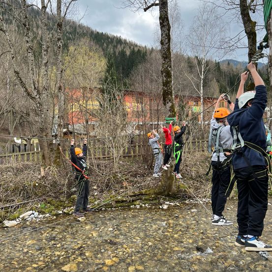 Eine Gruppe von Menschen nimmt an einer Seilrutschen-Aktivität teil. Sie tragen Sicherheitsausrüstung, darunter Helme, und sind mit einem Seil verbunden. Die Umgebung ist von Bäumen und einem Holzzaun umgeben, wobei ein Gebäude im Hintergrund zu sehen ist.
