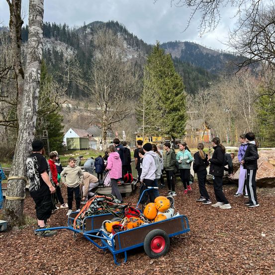 Eine Gruppe von Menschen steht in einem bewaldeten Gebiet mit Bergen im Hintergrund. Eine Person trägt ein schwarz-weißes Shirt. Ein blauer Wagen mit Kürbissen und Helmen steht vor der Gruppe.