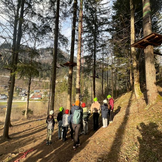 Eine Gruppe von Menschen befindet sich im Wald mit Bäumen und Häusern im Hintergrund. Sie tragen Helme und Rucksäcke. Sie betrachten die Plattform mit Seilen darüber.