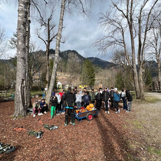 Eine Gruppe von Kindern steht draußen in einem bewaldeten Gebiet, einige sitzen auf dem Boden. Ein Mann mit einer schwarzen Jacke schiebt einen Karren mit Kürbissen.