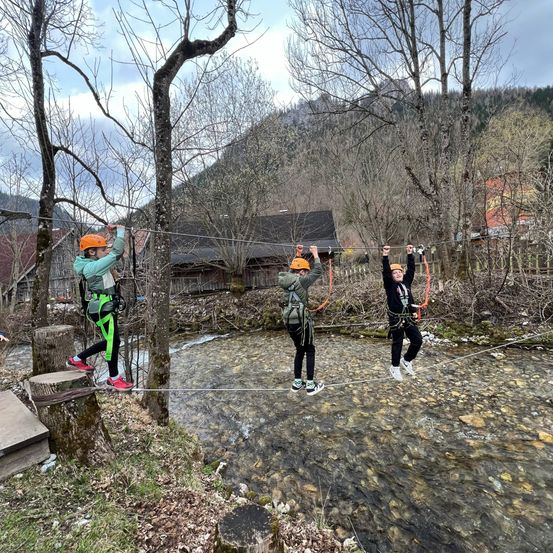 Drei Personen überqueren eine Hängebrücke über einen Bach. Sie tragen Sicherheitsausrüstung, darunter Helme und Gurte. Sie halten sich an Seilen fest. Die Brücke ist zwischen Bäumen errichtet.