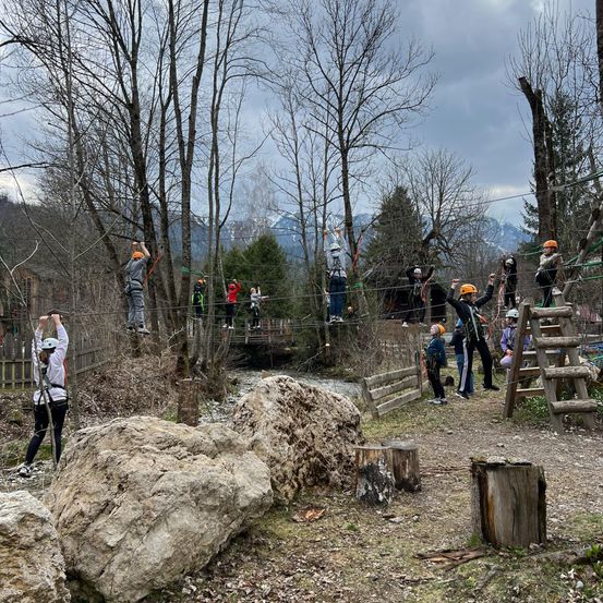 Eine Gruppe von Menschen überquert eine Seilbrücke über einem Fluss, mit Bergen im Hintergrund.
