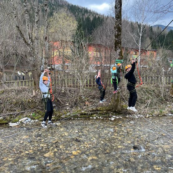 Vier Personen überqueren eine Hängebrücke über einem Bach. Sie tragen Helme und halten sich an den Sicherheitsseilen fest.