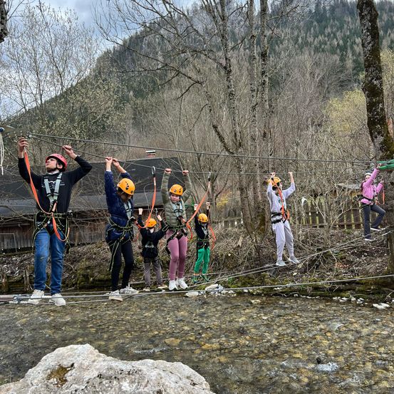 Eine Gruppe von Menschen mit Helmen und Sicherheitsgurten überquert eine Hängebrücke über einem Fluss, mit einem Berg im Hintergrund.