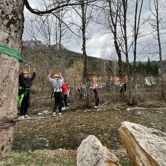 Eine Gruppe von Menschen mit Helmen und Turnschuhen überquert mit einer Seilbahn einen Bach im Wald.